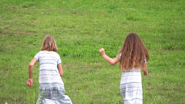 Slow motion. Two sisters teenagers in long sundresses run on green grass.