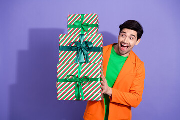 Obraz premium Man in orange jacket holding a tall stack of striped, ribbon-wrapped Christmas presents with a surprised happy expression on purple background
