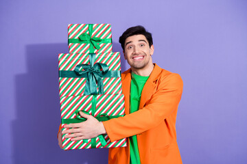 Obraz premium Smiling man holding stack of wrapped Christmas presents with green ribbons against purple background, festive holiday gift giving and celebration portrait in bright orange jacket