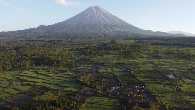 Drone view of Mount Semeru overlooking the agricultural landscape of Pronojiwo, East Java, with green farmland and volcanic scenery illuminated by morning light.