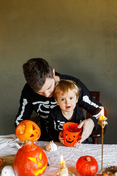 Dad and son in skeleton costumes making jack o lantern together. Happy Halloween. Father with child carving pumpkins on table with spooky decor and candles on dark background. Space for text