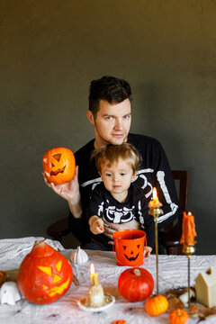 Dad and son in skeleton costumes making jack o lantern together. Happy Halloween. Father with child carving pumpkins on table with spooky decor and candles on dark background. Space for text