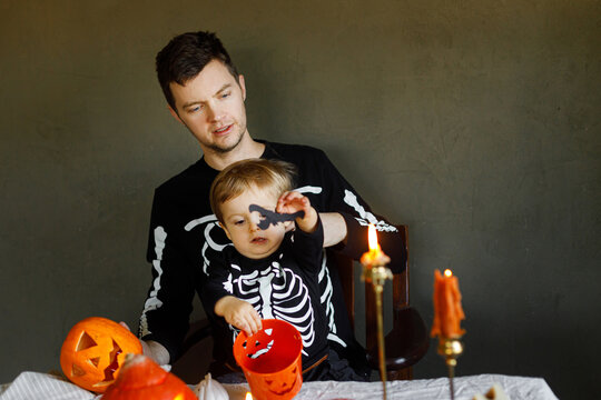 Dad and son in skeleton costumes making jack o lantern together and playing with bucket trick or treat. Happy Halloween. Father with child carving pumpkins on table with spooky decor and candles