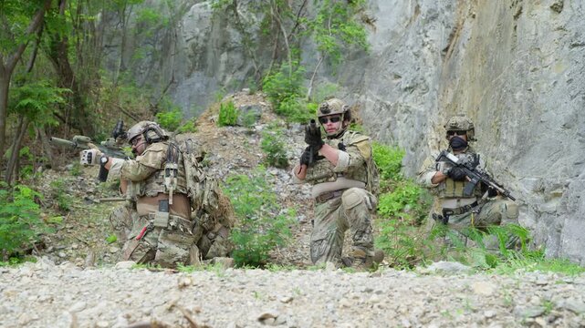 Squad of five soldiers in camouflage uniform kneeling and aiming rifles during military tactical operation outdoor, special forces operators holding weapons near rock cliff