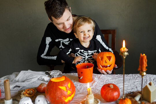 Happy Halloween. Father with child carving halloween pumpkin on table, making scary decorations on dark background. Space for text. Dad and son in skeleton costumes cutting jack o lantern together