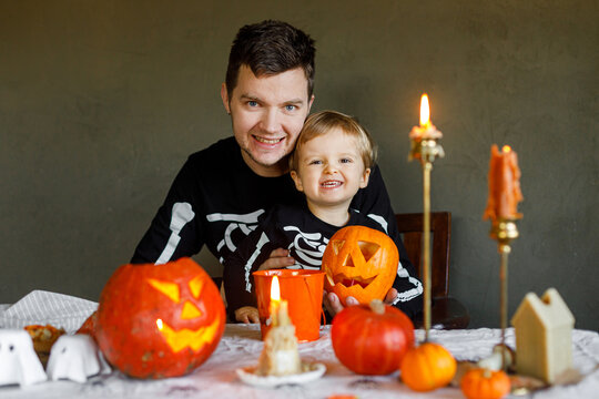 Dad and son in skeleton costumes making jack o lantern together and smiling. Happy Halloween. Father with child carving pumpkins on table with spooky decor on dark background. Space for text