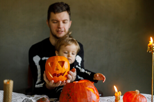 Happy Halloween. Father with child carving halloween pumpkin on table, making scary decorations on dark background. Space for text. Dad and son in skeleton costumes cutting jack o lantern together