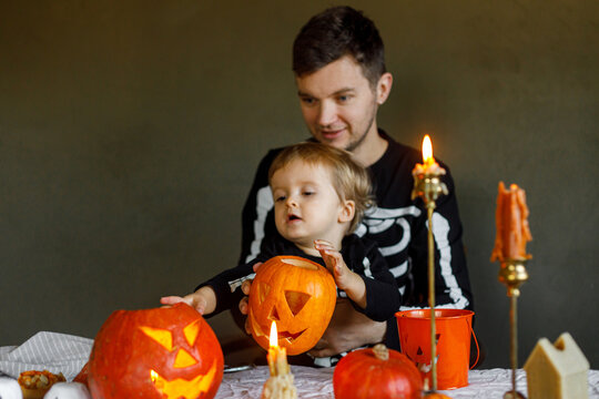 Dad and son in skeleton costumes making jack o lantern together. Happy Halloween. Father with child carving pumpkins on table with spooky decor and candles on dark background. Space for text