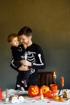 Dad and son in skeleton costumes standing at table with spooky decor and candles on dark background. Happy Halloween. Father with child carving pumpkins