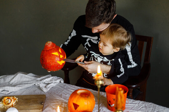 Happy Halloween. Father with child carving halloween pumpkin on table, making scary decorations on dark background. Space for text. Dad and son in skeleton costumes cutting jack o lantern together