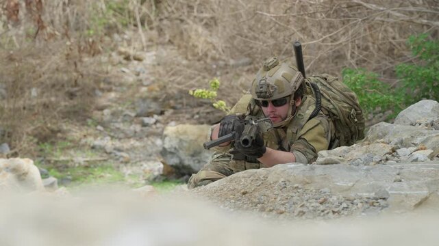 Soldier in camouflage uniform lying in prone position aiming rifle behind rocks. Tactical military warrior holding weapon during combat training outdoor