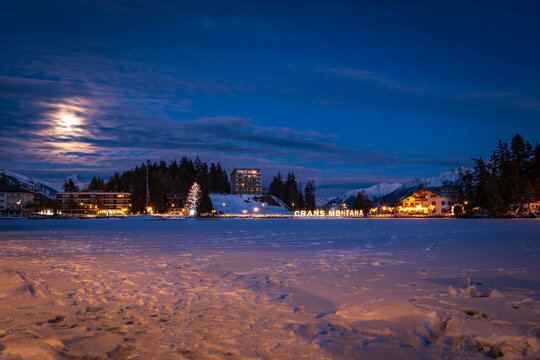 Scenic view of frozen and snow covered Etang Grenon (Lac Grenon) with illuminated lettering "Crans Montana", Switzerland against mountains and blue sky with clouds in the evening