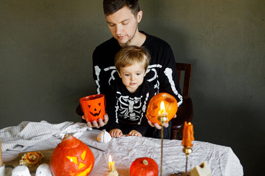 Happy Halloween. Father with child carving halloween pumpkin on table, making scary decorations on dark background. Space for text. Dad and son in skeleton costumes cutting jack o lantern together