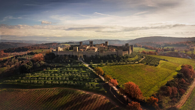An aerial view of a medieval walled village perched on a Tuscan hill, surrounded by golden vineyards and olive groves during a serene late afternoon sunset in Italy.