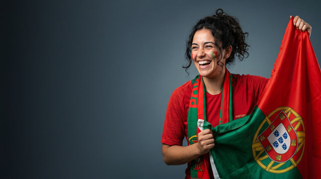 Cheerful Portugal football fan holding national flag, happy female supporter in red jersey with face paint laughing, cheering for Portuguese soccer victory isolated on studio background