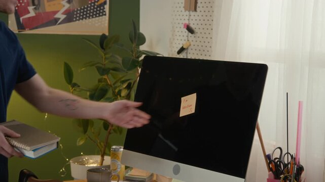 Young Caucasian male student walking to desk, removing sticky note from broken computer in shared dorm room, looking angry and frustrated, representing gen Z financial struggles and roommate conflictz