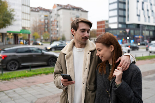 A happy young couple checks a smartphone while walking together on an urban sidewalk.