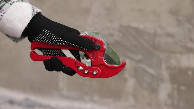 A hand in a work glove holds a red pipe cutter for plastic pipes. A hand holds professional scissors for cutting PVC and polypropylene pipes. A red tool with a sharp steel blade