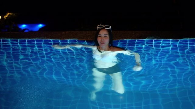 Happy Asian woman turning and swimming in a glowing blue illuminated swimming pool at night