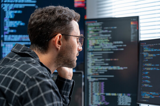 Male data scientist and software engineer code on multiple screens in a modern hybrid office building machine learning models optimizing databases and implementing AI driven solutions in daytime.