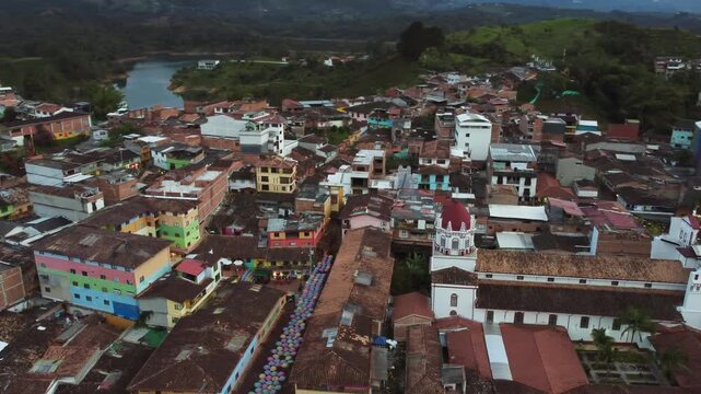Guatap&eacute;, Antioquia - Aerial Orbit of colorful town buildings and church with El Pe&ntilde;&oacute;n de Guatap&eacute; rock in background on cloudy day
