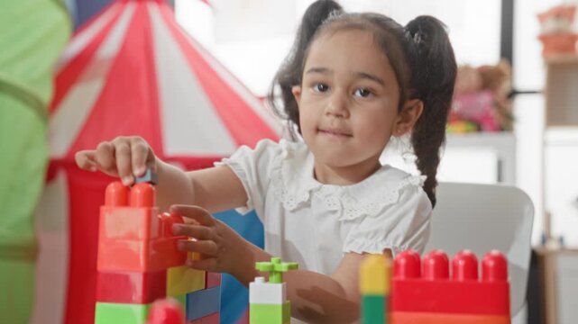 Girl stacking colorful plastic blocks with hand at a play table inside building; curiosity concentration focus.