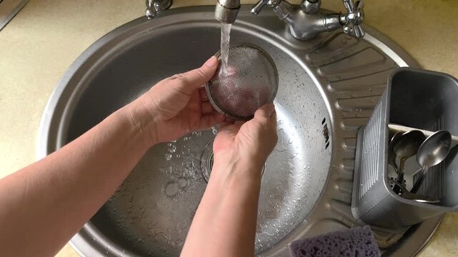 Person washes sieve in metal kitchen sink. Hands clean stainless steel sieve after ground coffee under water stream. Housework involves washing sieve carefully. Domestic routine includes cleaning.