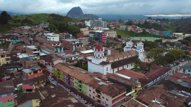 Aerial Overview of colorful buildings and church in Guatap&eacute;, Antioquia, with El Pe&ntilde;&oacute;n de Guatap&eacute; rock in background on cloudy day in 4k