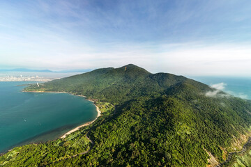 Naklejka premium An aerial view shows the Son Tra Peninsula in Da Nang, Vietnam, with its densely forested mountain slopes meeting the clear blue ocean and a sandy beach