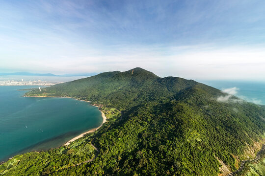 An aerial view shows the Son Tra Peninsula in Da Nang, Vietnam, with its densely forested mountain slopes meeting the clear blue ocean and a sandy beach