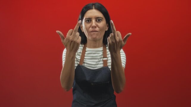 Woman wearing striped shirt and denim apron shows middle fingers with both hands and sticks out tongue in red studio; defiance.