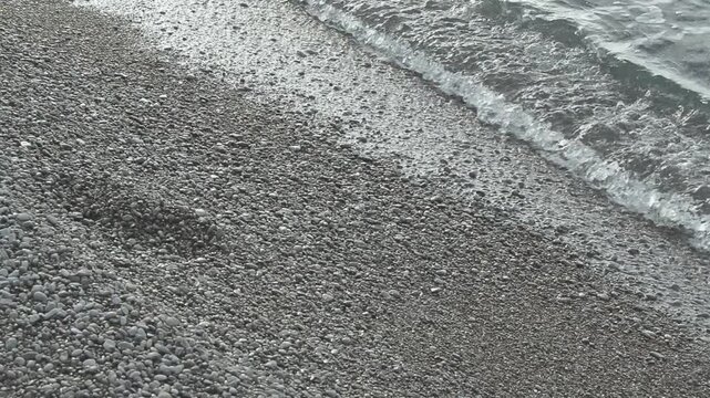 Gentle waves lap against a pebbled shore, revealing a gradual retreat of water, showcasing the rhythmic motion of nature along a tranquil beach setting