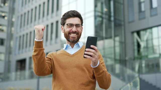 Happy excited businessman received great news on mobile phone standing on the street near modern office building. Smiling shocked surprised worker in glasses reads positive good message on smartphone