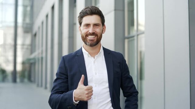Smiling businessman in formal suit showing thumb up looking at camera while standing on street near office building. Happy positive male shows gesture of like, agreement, success, approval. Close up