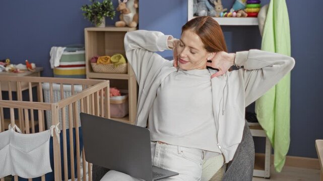 Woman stretches arms overhead beside laptop and crib in nursery while seated in chair and smiling softly; home work balance relaxed.