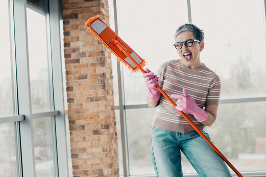 Joyful mature woman singing and dancing while cleaning with a mop and pink rubber gloves in a bright modern apartment