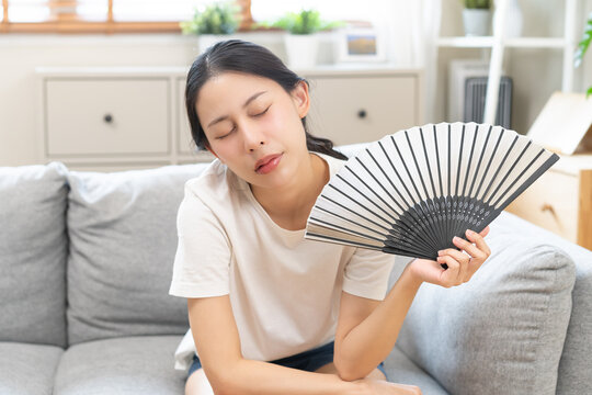 Suffering summer heat wave stroke, hot weather, tired asian young woman feeling discomfort sweaty, sitting on couch in living room, hand fan in blowing, wave fan to ventilation when high temperature.