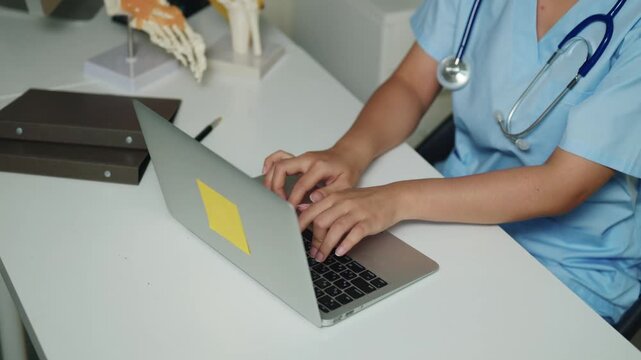 Doctor in Blue Scrubs Working on Laptop at Medical Desk with Stethoscope and Bone Model