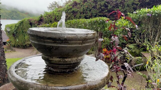 static shot of a rustic stone water fountain in a lush garden overlooking a lake