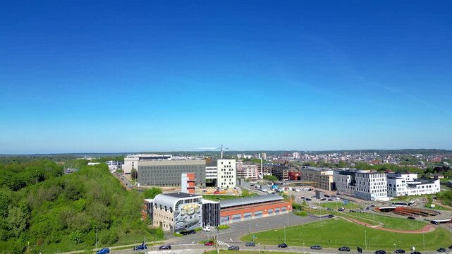 Cinematic aerial view of modern Leuven, Belgium, featuring the Gasthuisberg university hospital campus and fire station under a bright blue sky.