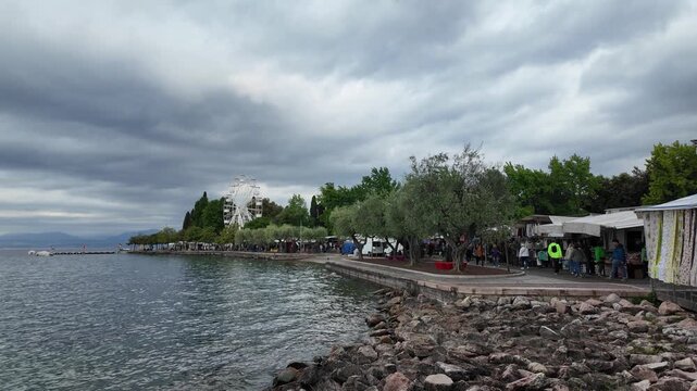 Bardolino, Italy - May 14, 2026 - Lakeside promenade with market stalls and Ferris wheel under cloudy skies