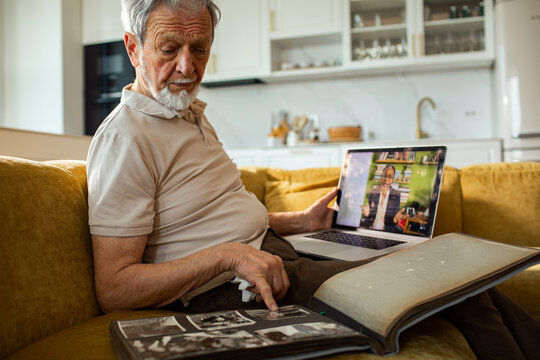 Senior man reminiscing with photo album during video call at home