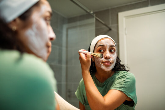 Young woman applying facial mask with brush in bathroom mirror