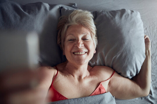 Happy senior woman taking selfie in bed at home