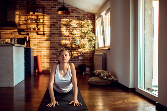 Young woman practicing cobra yoga pose at home in sunlit living room