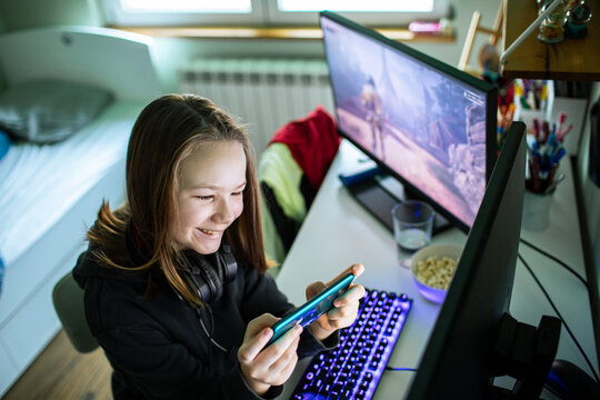 Smiling teen girl playing mobile game at home computer desk