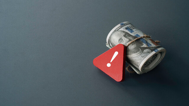 A rolled bundle of US dollar bills tied with twine next to a red exclamation mark warning icon depicting financial danger and economic risk.
