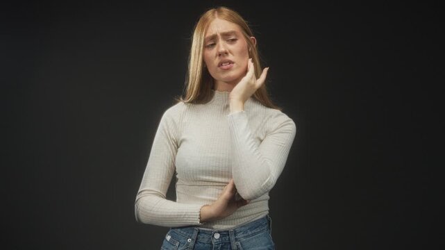 Woman touching ear with hand and holding cheek in studio while wearing turtleneck and jeans, closed eyes and pained grimace; earache pain.