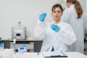 Fototapeta premium Expert professional Caucasian female scientist wearing safety glasses and blue gloves using a glass dropper for chemical liquid analysis in a modern biotechnology pharmaceutical laboratory.