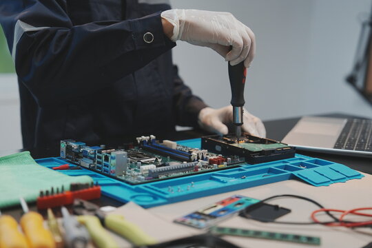 A technician wearing white gloves uses a screwdriver to assemble or repair a hard drive placed on a blue silicone work mat. In the background, a computer motherboard 